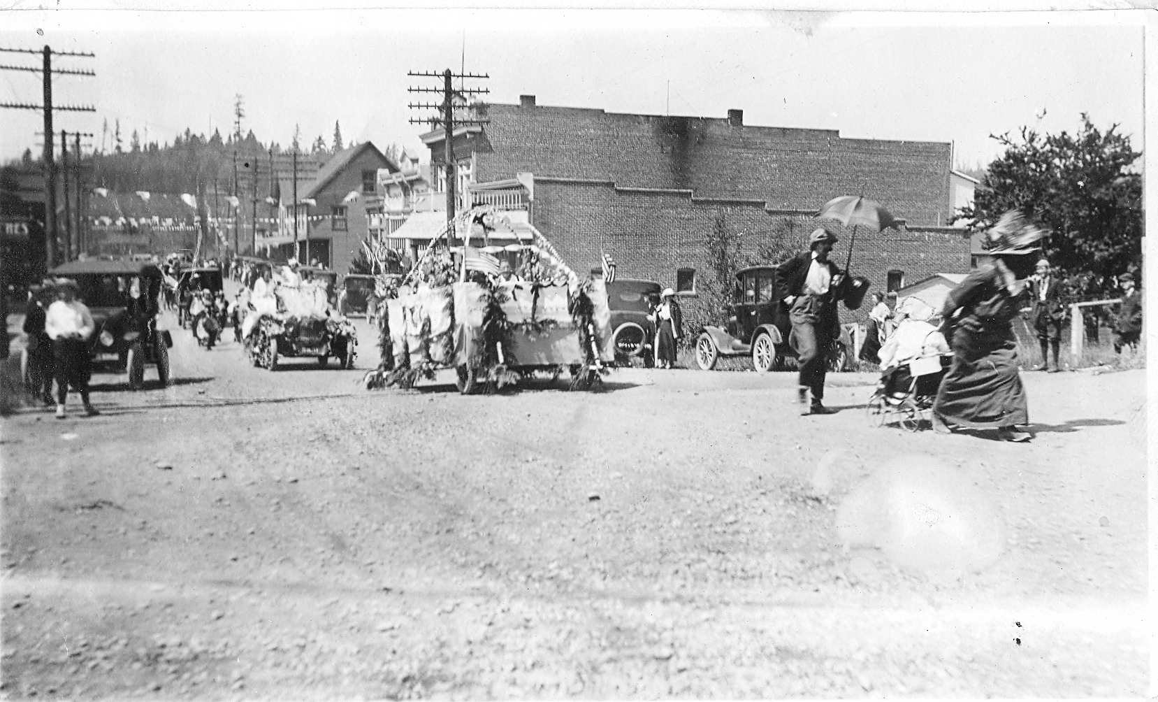 Handwritten note on paper attached to photo states "July Parade - Bothell - 1915." The view is looking west on Main Street. A woman in a hat and pulling a baby carriage is walking in front of the parade, followed by a man with a parasol. A float is at the front of the parade followed by a car carrying women dressed in white, then people marching, other cars, and people on horseback. Dexter's Cafe and Mohn Furniture are in the brick buildings on the right.