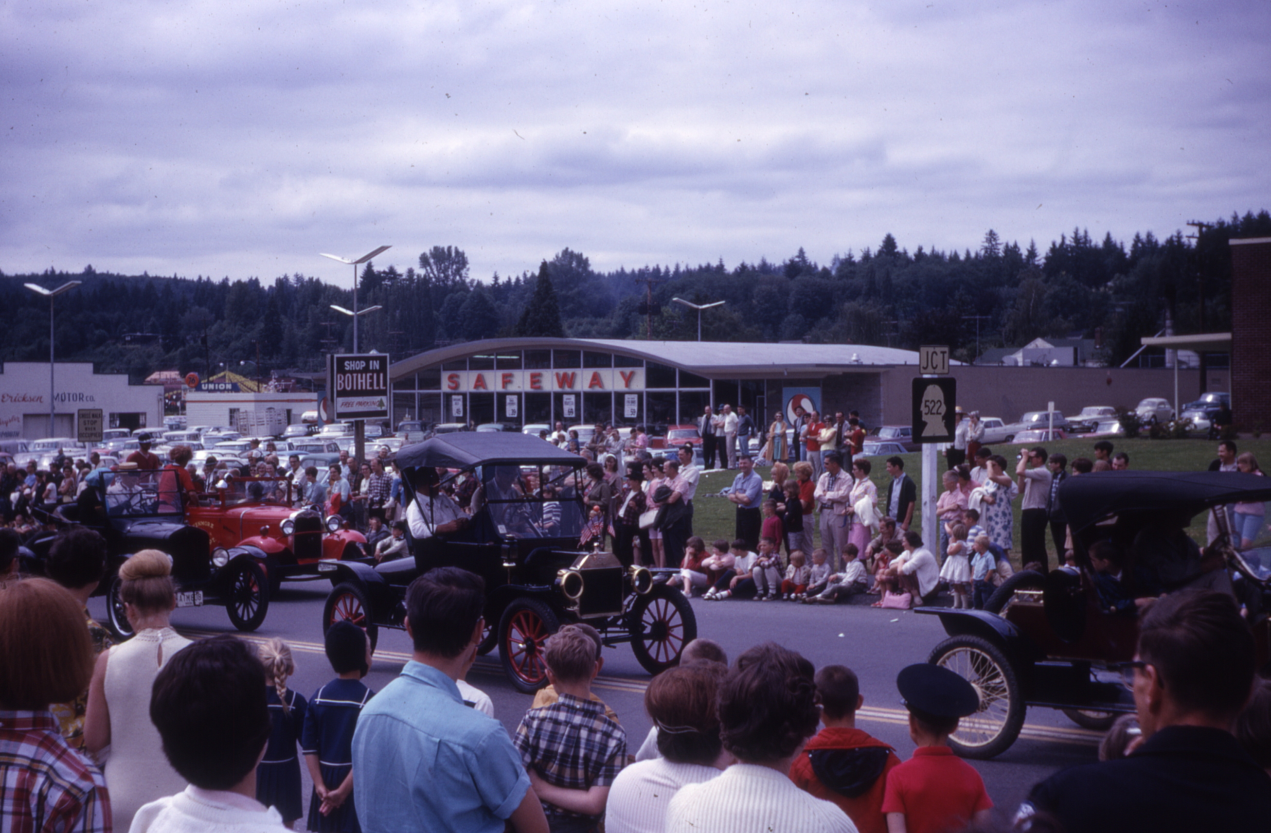 The parade route turned north from Main Street on the Bothell-Everett Highway, past the new Safeway (built in 1962 on the site of the Avon Theater). Many classic cars driven by members of the Horseless Carriage Club participated in the parade. In the background to the left can be seen Ericksen Motor Company, a Chrysler-Plymouth dealership owned by third-generation Bothell merchant Bud Ericksen, and the Union 76 service station owned by Lowell Haynes.