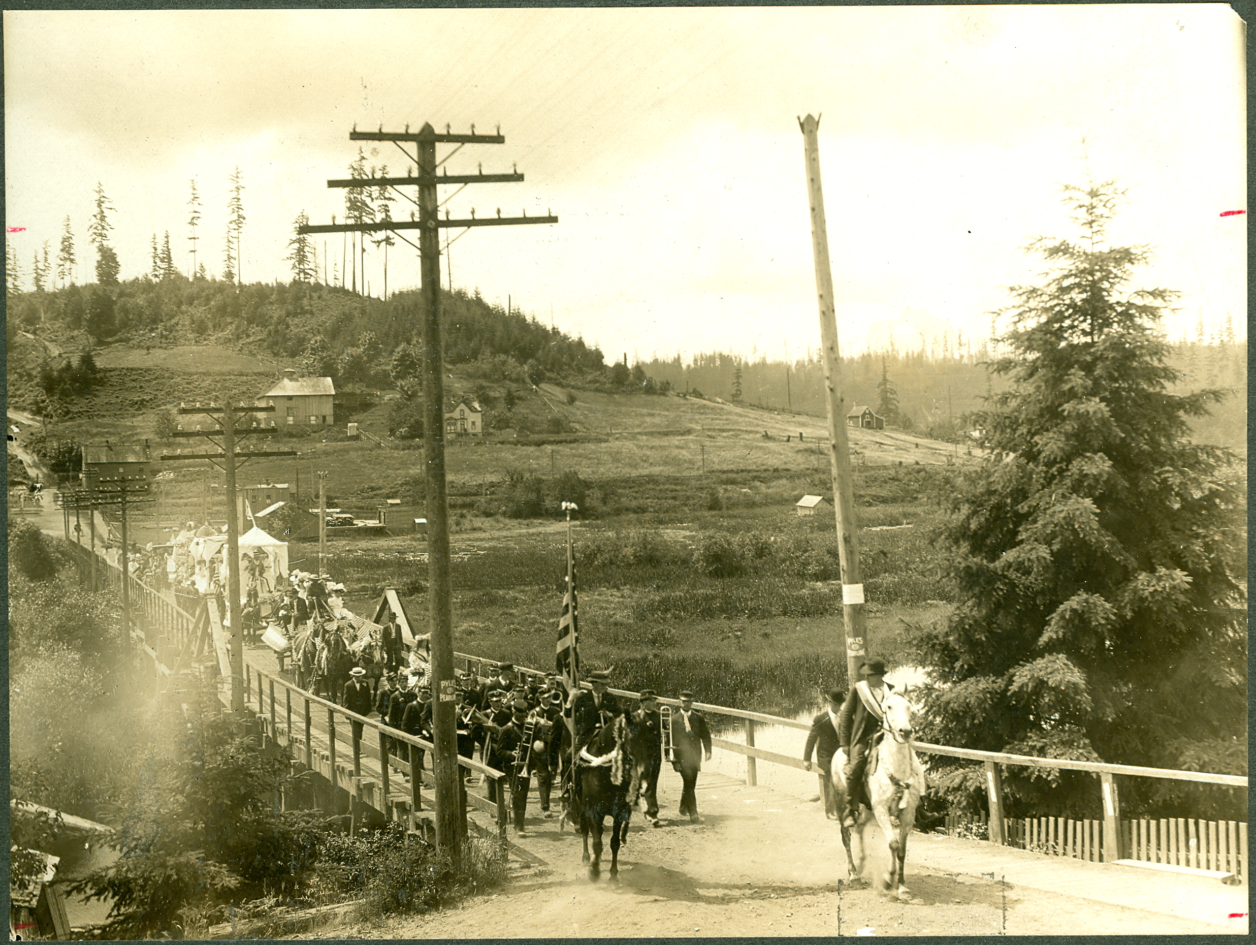 Fourth of July Parade, probably 1907, possibly 1908. On the back of a copy of the photo is typed, "Shows the Bothell Town Band leading a parade back across the Riverside Bridge toward Main Street." Several floats are following the band. A man with a sash mounted on a light-colored horse leads the parade, followed by a male flag bearer mounted on a dark horse. The poles on either side of the bridge in the foreground have signs pasted on them that read Pyles Pearline, which was an advertisement for a washing compound. Norway Hill is to the immediate right side of the bridge. Handwritten on the back of one of the original photos is, "House straight back from end of bridge was home of my grandmother Ida Kraase and her daughter Bertha and (her son ??) -in-law Robert John Magwood for many years. In center is old Pearson home. My father lived there ... little over a year and died there September 17, 1953. Beatrice Spinney McGarrigle." We believe the dark house closest to the road at the end of the bridge is the home of Ida Kraase. Squak Slough by Amy Stickney, says the photo probably taken in 1907. A copy of the photo says that it was taken prior to 1908. Handwritten in pencil on the back of one of the original photos is "1908." Handwritten on the back of one of the original photos is, "Donated by Beatrice McGarrigle 10/28/72." On the back of the copy of the photo is typed, "Photo loaned by Mrs. Ross Worley Sr., Woodinville." 