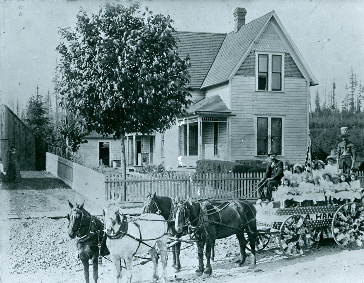 The W. A. Hannan float waiting in front of the W. A. Hannan house on Bothell's Main Street. Mr. Hannan operated a general store on Main Street, so the float represented his business. The people are unidentified, but it may be that W.A. Hannan himself is the driver (teamster), and his daughter Gladys may be seated just below his left side. Note the Uncle Sam costume on the man standing at the back of the wagon.